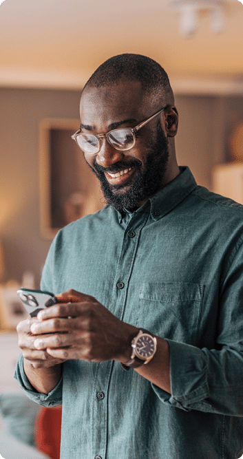 A person wearing glasses and a green button-up shirt is smiling while looking at a smartphone in a warmly lit room. Description generated by AI